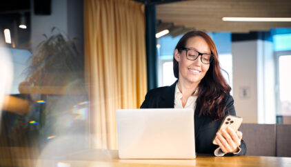 A smiling woman in a modern office. Working from the office, a business woman in eyeglasses using a smart phone and a laptop. Multi-tasking concept and copy space for your design.