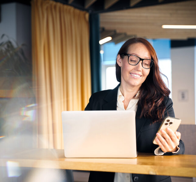 A smiling woman in a modern office. Working from the office, a business woman in eyeglasses using a smart phone and a laptop. Multi-tasking concept and copy space for your design.